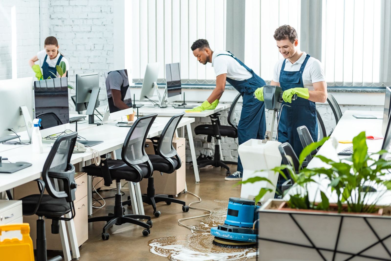 Commercial cleaning crew in gloves wiping desks and scrubbing the floor of an open office