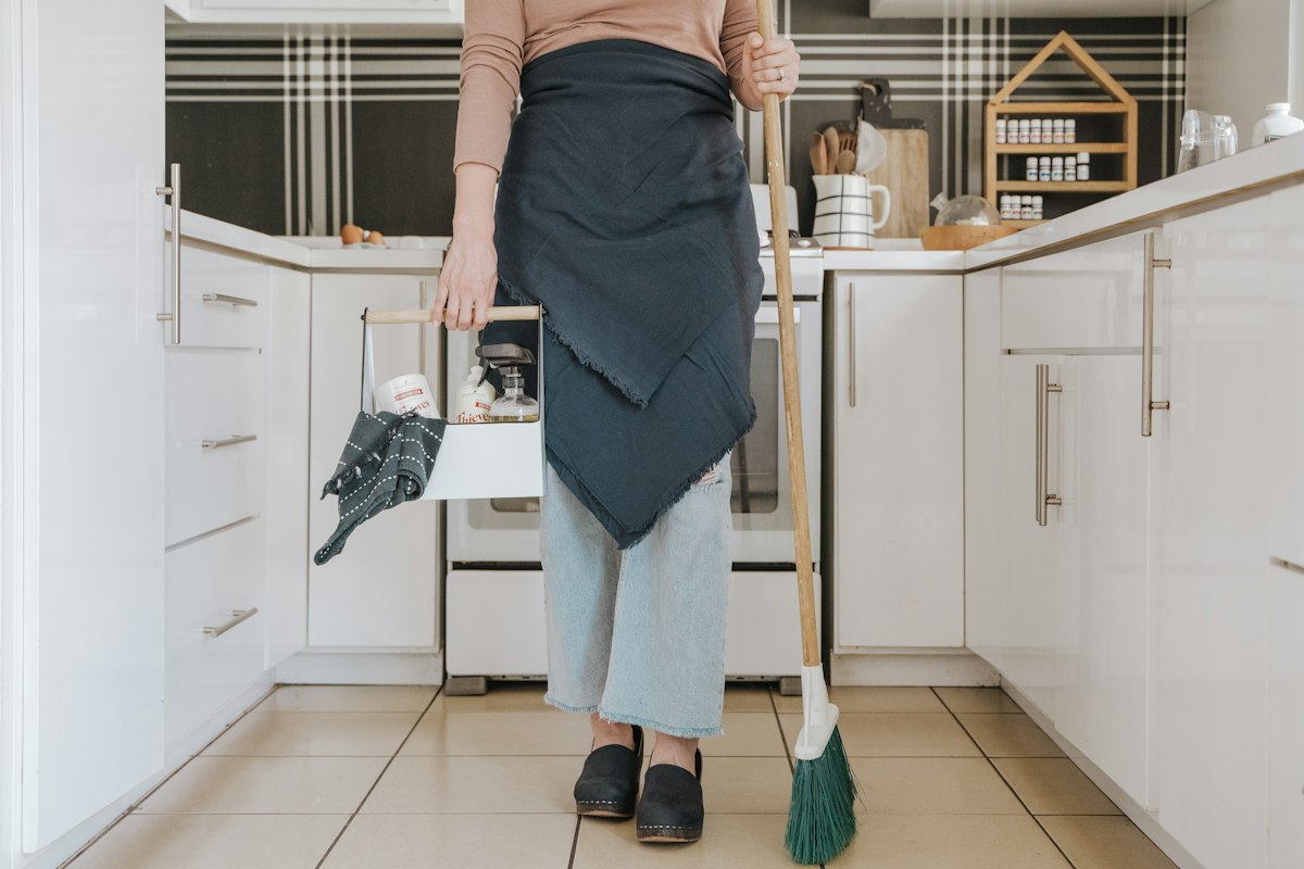 Housekeeper with a broom and cleaning caddy in a bright white kitchen in Northern Virginia